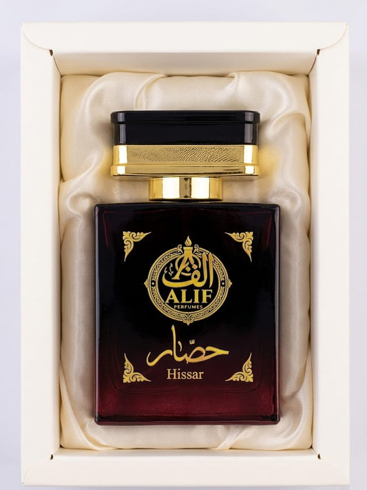 A bottle of Alif Hissar perfume presented in a cream-colored gift box with cream satin lining. The bottle is dark brown with gold lettering and a gold cap.