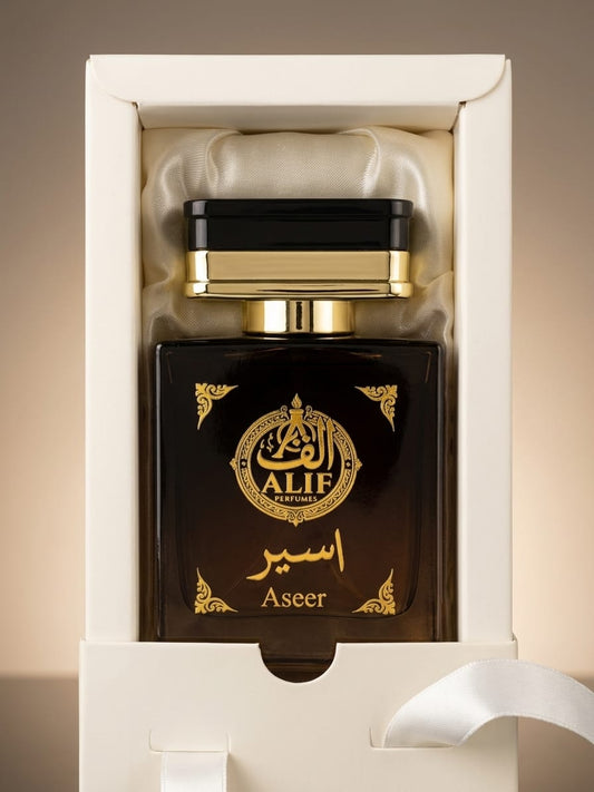 A luxury perfume bottle labeled 'ALIF Aseer' in elegant gold Arabic calligraphy, presented inside an open white gift box with a satin lining.