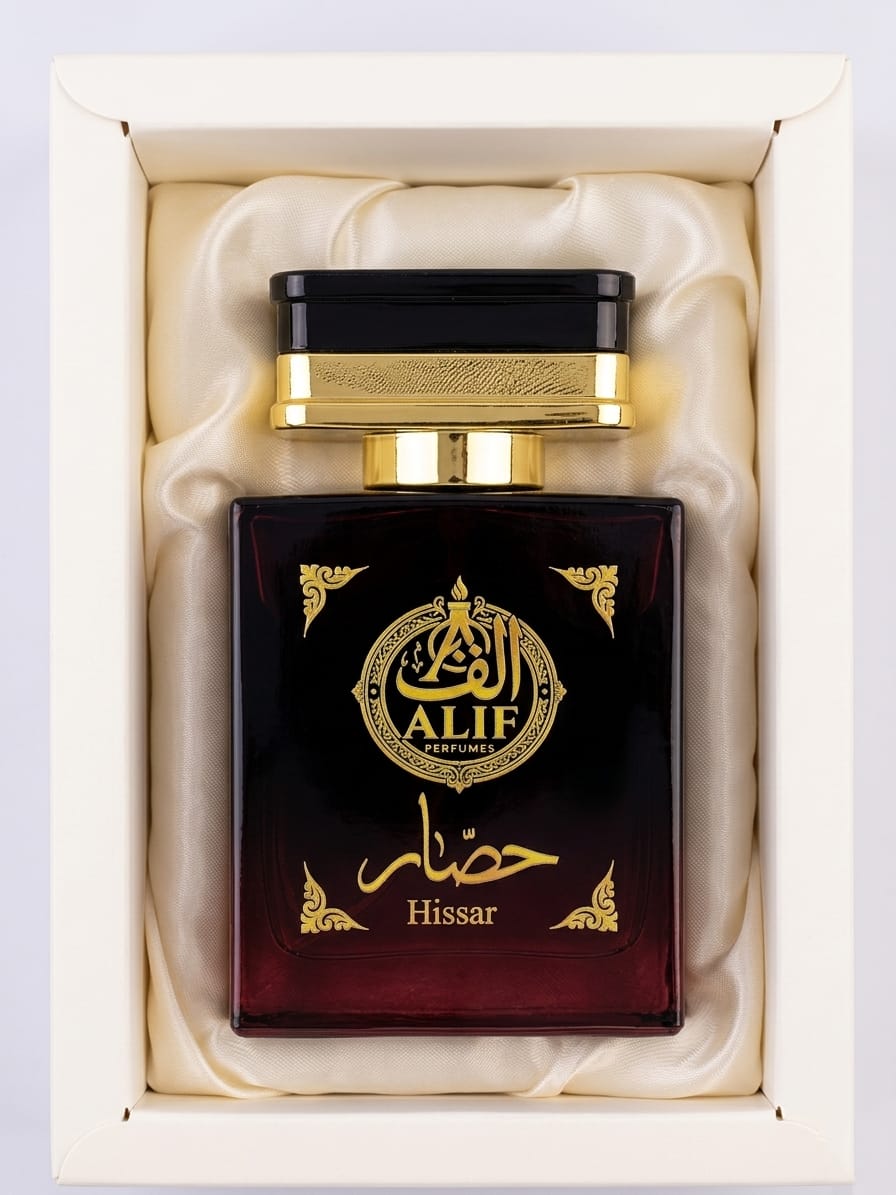 A bottle of Alif Hissar perfume presented in a cream-colored gift box with cream satin lining. The bottle is dark brown with gold lettering and a gold cap.