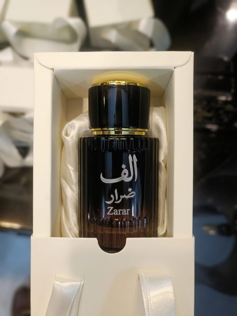 A dark amber glass perfume bottle with a black cap and gold trim, displayed inside an open white gift box with satin ribbon. The bottle features Arabic calligraphy and the brand name 'Alif' Zarar printed in white and silver.